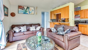Een woonkamer met twee bruine leren banken, een glazen salontafel en een keuken met houten kasten en apparatuur.