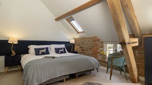 Un dormitorio con techo inclinado, una cama con almohadillas estampadas, una mesita de noche, una silla y una pared de piedra.