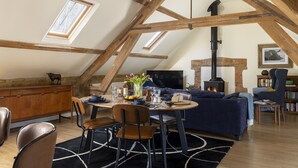 Un espacio tipo loft con vigas de madera a la vista, una chimenea, una zona de comedor con mesa y sillas, y un rincón de descanso acogedor con un sofá y sillones.