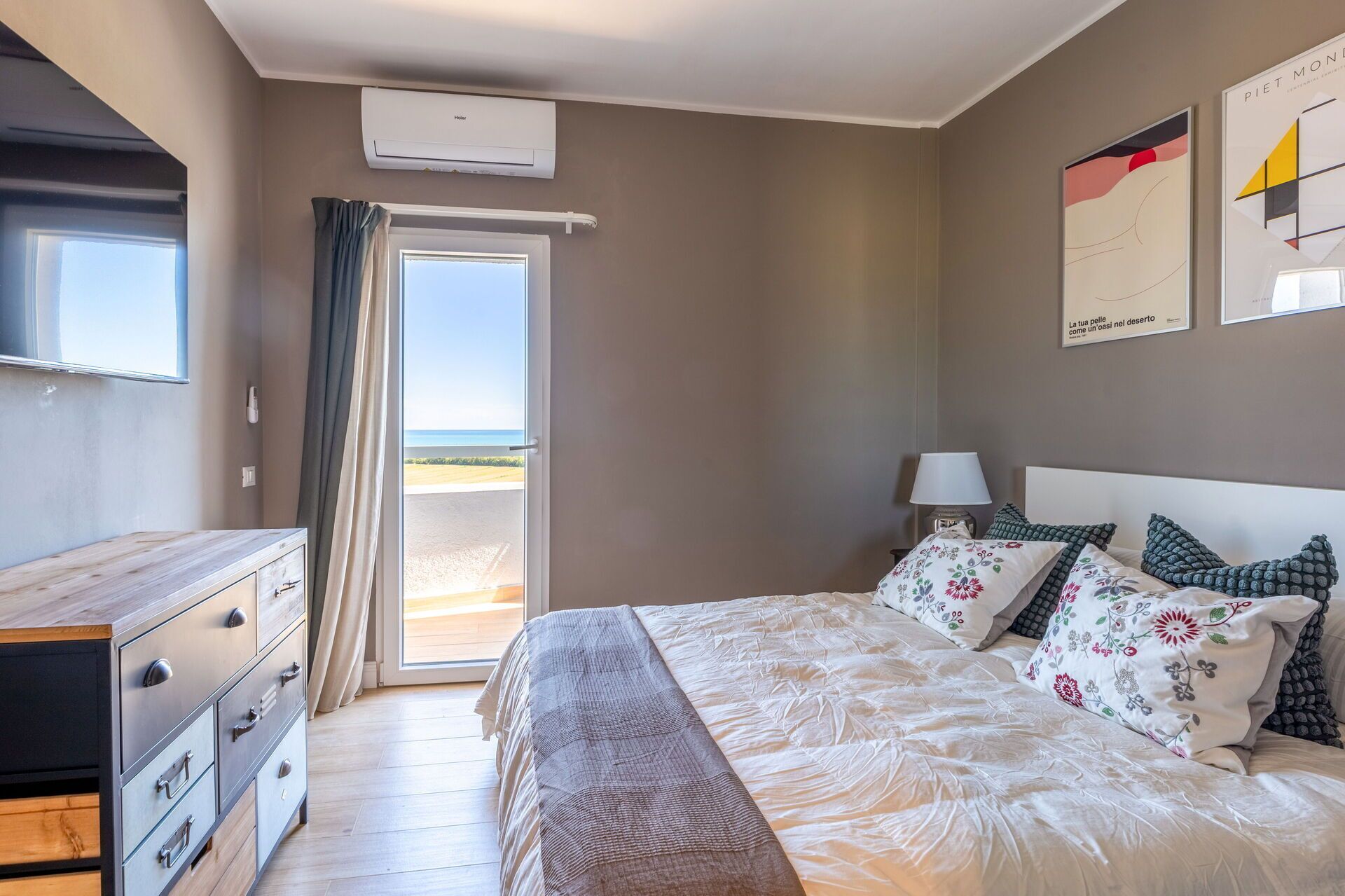 A bedroom with a bed, a dresser, a nightstand, and a window with a view of the beach.
