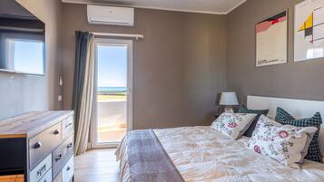 A bedroom with a bed, a dresser, a nightstand, and a window with a view of the beach.