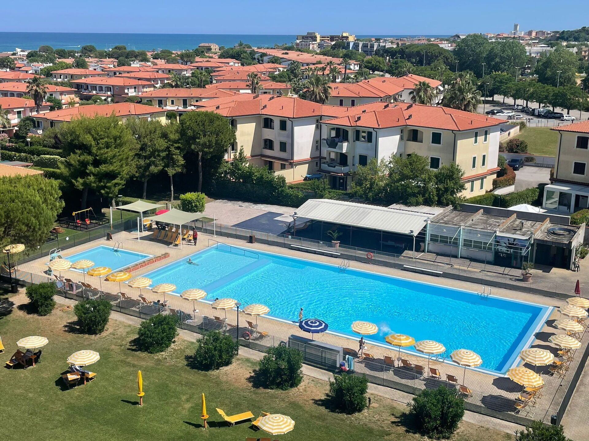 Una gran piscina con múltiples carriles, rodeada de tumbonas y sombrillas, con edificios residenciales y un cielo despejado de fondo.