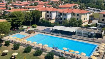 Una gran piscina con múltiples carriles, rodeada de tumbonas y sombrillas, con edificios residenciales y un cielo despejado de fondo.