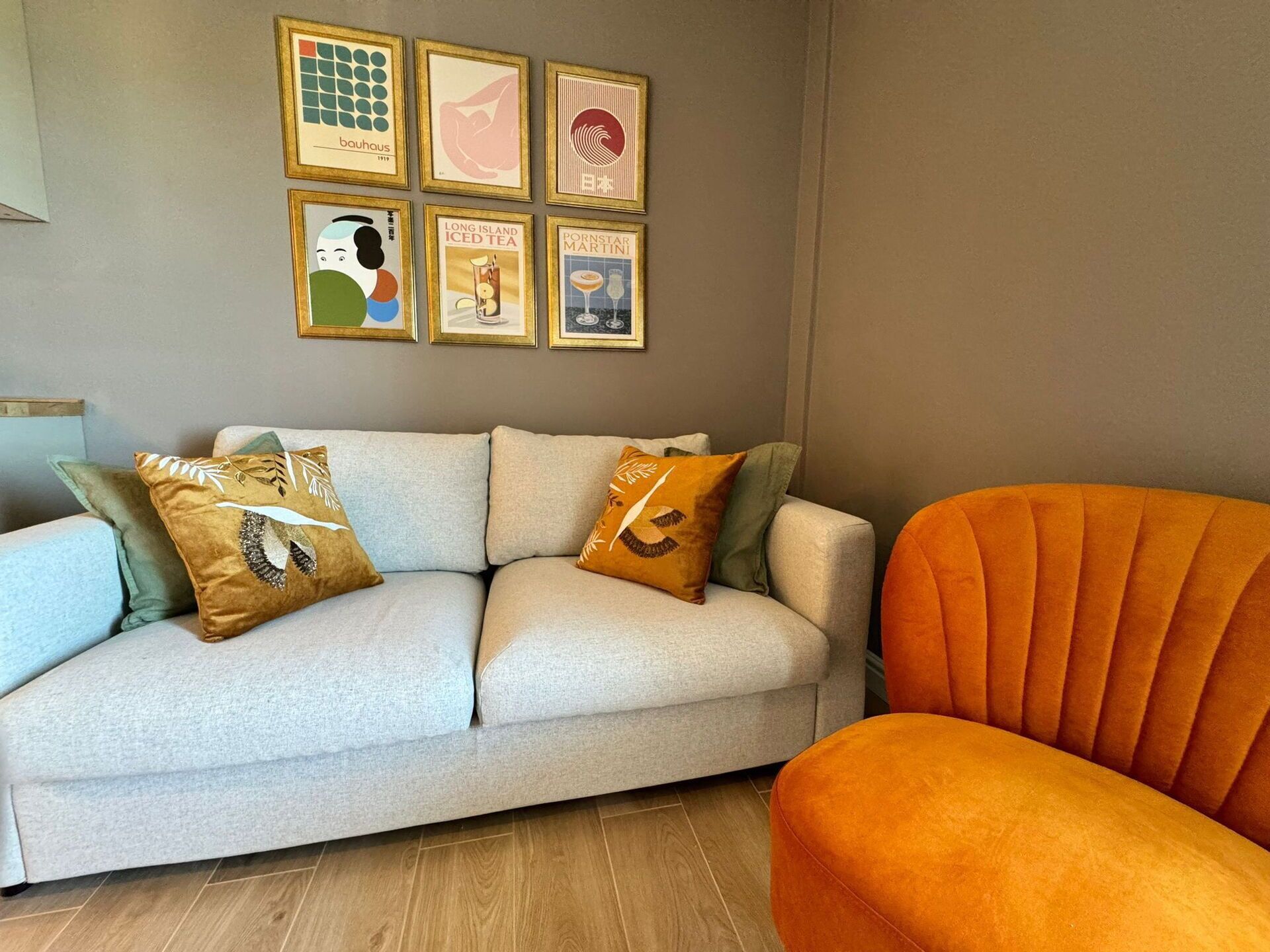 A living room with a light grey sofa, orange armchair, and six framed artworks on the wall.