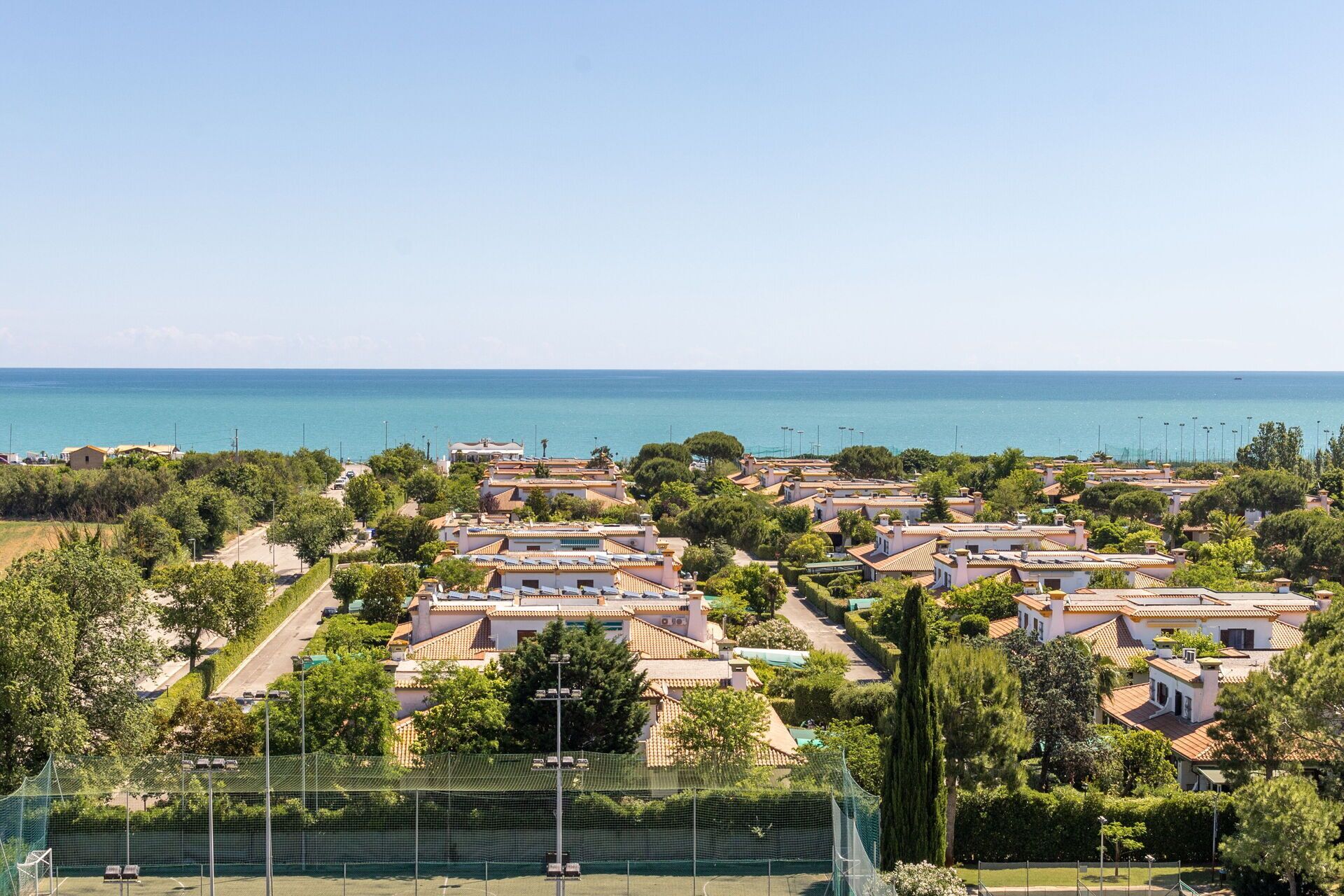 A coastal residential area with terracotta-roofed houses, a tennis court, and a clear view of the sea.