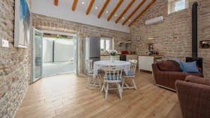 A spacious living area with a stone wall, wooden beams, a dining table with chairs, a sofa, and a kitchen area with a stove. - Villa Pod Murvom (Novigrad)