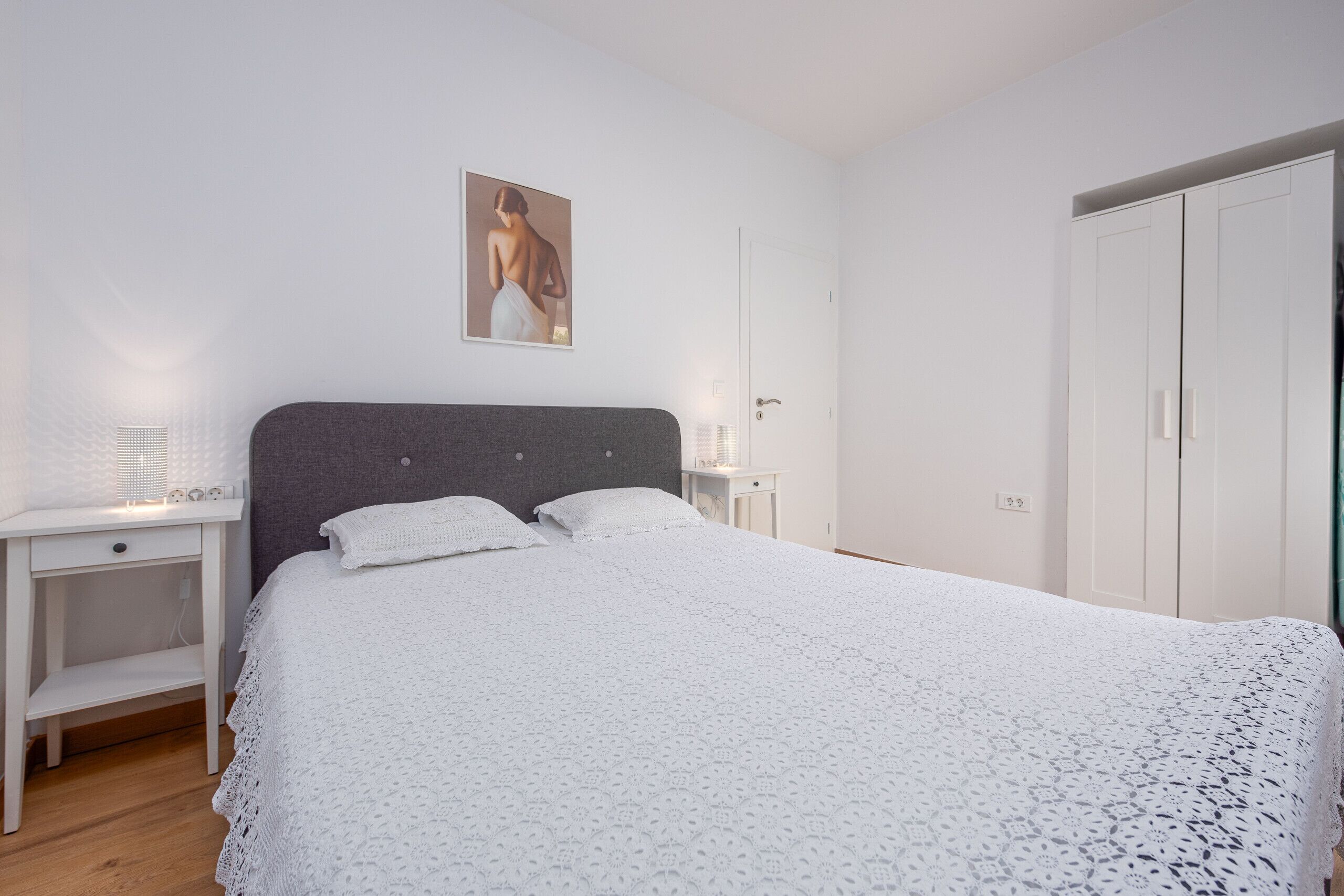 A neatly arranged bedroom with a large bed, a bedside table with a lamp, a wardrobe, and a framed picture on the wall.