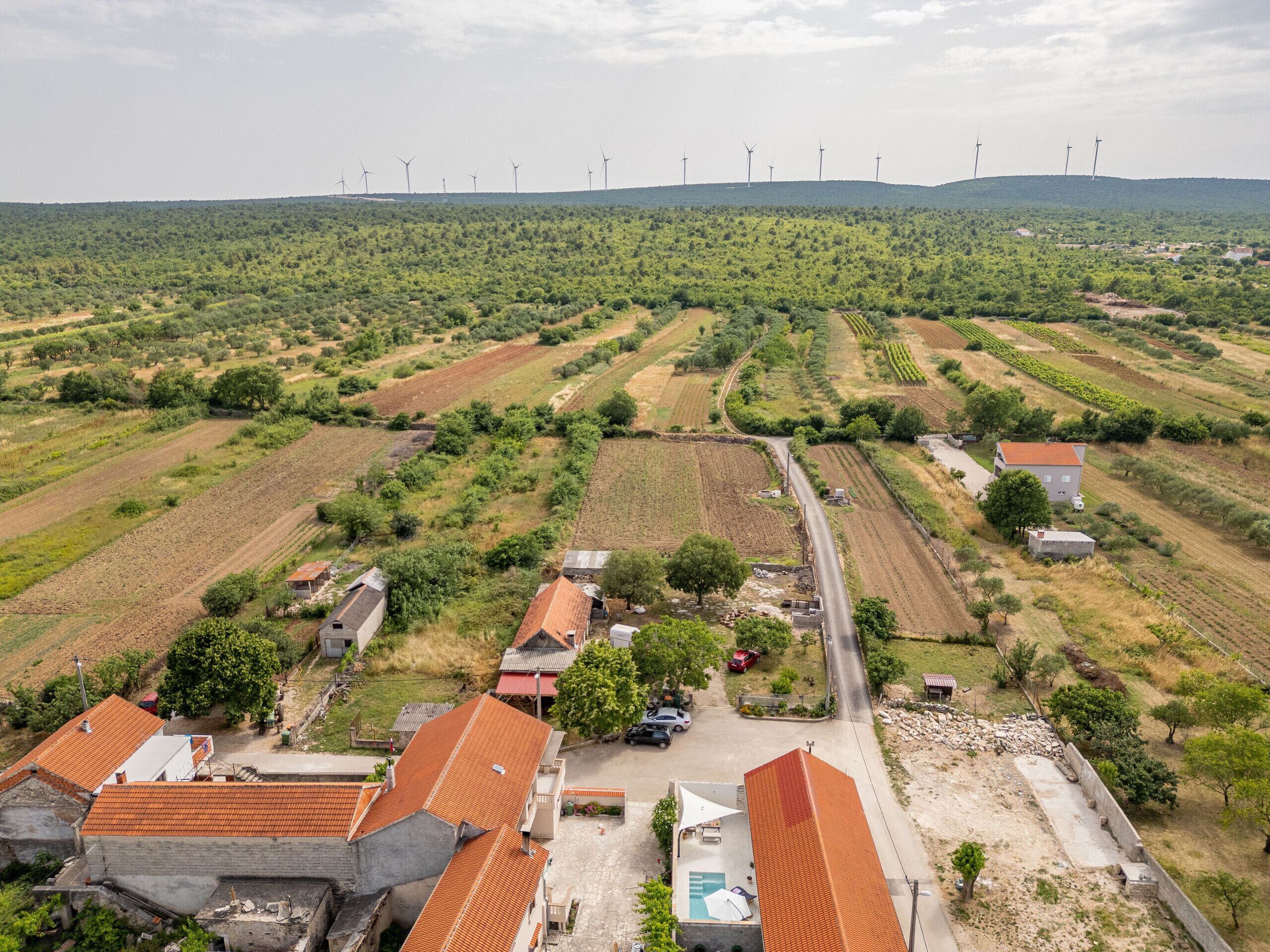 An aerial view of a rural area with wind turbines in the background, agricultural fields, and scattered houses with red-tiled roofs.