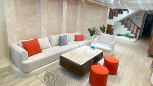 A modern living room with a sectional sofa, a marble coffee table, and two red ottomans. A staircase is visible in the background. - CC Xanh Xanh (Xuan Huong - Da Lat)
