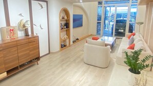 A modern living room with a wooden cabinet, a white sofa, and a large window offering a view of the outside. - CC Xanh Xanh (Xuan Huong - Da Lat)