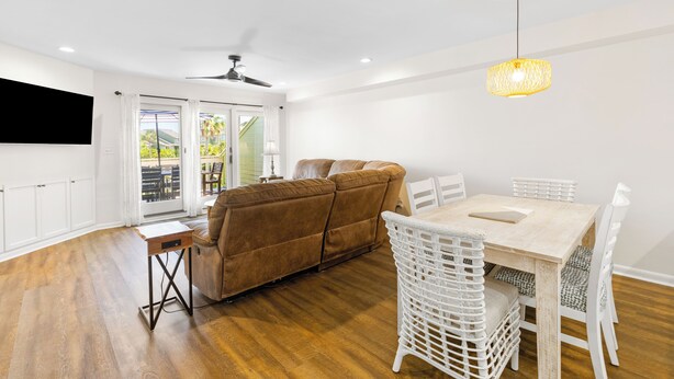 A modern living room with a brown sofa, white wicker chairs, a wooden dining table, and a ceiling fan.