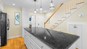A kitchen with a granite countertop, white cabinets, a black refrigerator, pendant lights, and a wooden staircase. - Beach Club Villa 38 - Oceanfront Gem! Walkout! (Isle of Palms)