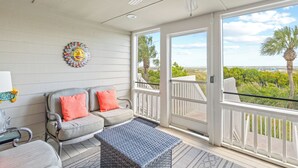 A spacious balcony with a view of the ocean and palm trees, a wicker sofa with orange cushions, and a ceiling fan. - Beach Club Villa 38 - Oceanfront Gem! Walkout! (Isle of Palms)