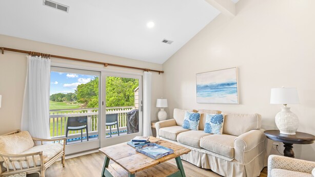 A spacious living room with a wooden coffee table, a beige sofa, and a view of a lush green landscape through large sliding glass doors. - Fairway Dunes 14 - Tennis! Pool! Beach! (Isle of Palms)
