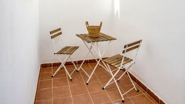 Un patio con paredes blancas, detalles en amarillo y piso de baldosas. Hay dos sillas plegables y una mesa pequeña con una cesta.