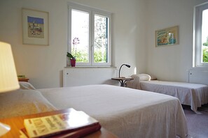 A room with two single beds, a desk with a lamp, and a window with a view of greenery. - Villa Flores a Mondello (Palermo)