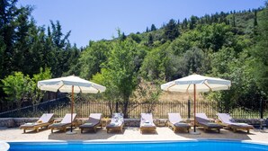 Zona junto a la piscina con sillas de descanso y sombrillas, rodeada por una valla y árboles.