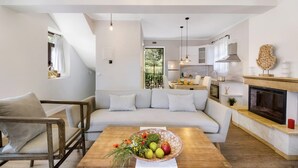 Una sala de estar moderna con un sofá, una mesa de centro de madera con fruta y flores, una chimenea y una cocina al fondo.