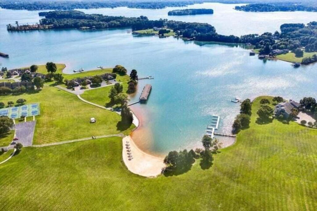 An aerial view of a lakeside property with a dock, a sandy beach, and surrounding greenery.