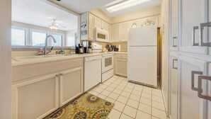 Una cocina compacta con armarios blancos, microondas, refrigerador, lavavajillas y una ventana con persianas.