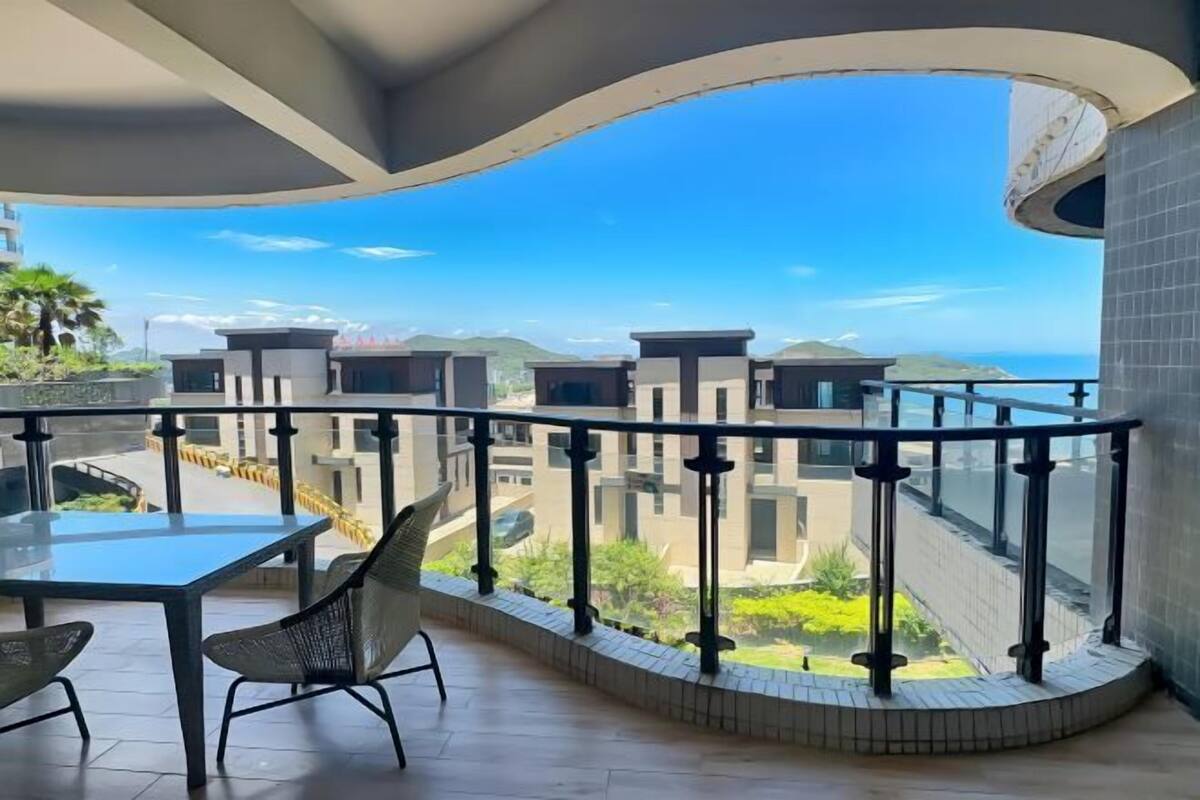 Un balcón con una mesa y sillas de vidrio, barandilla curva y vista a edificios modernos y follaje.