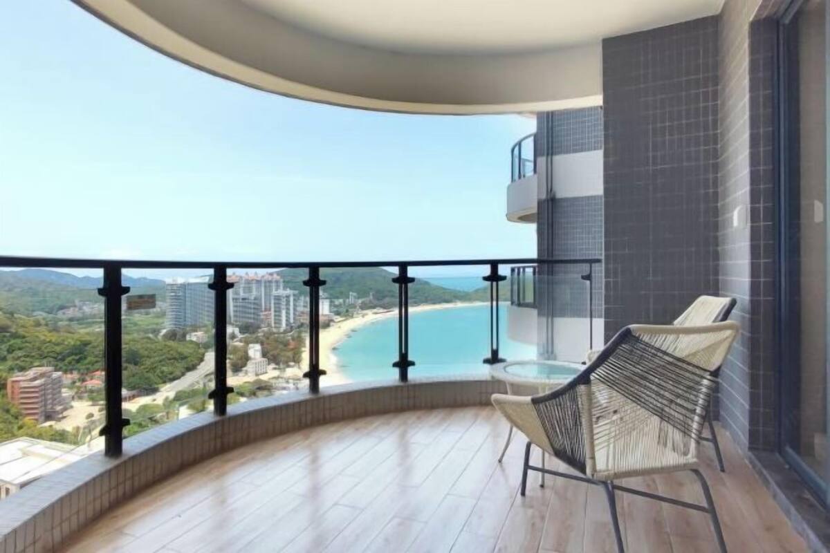 Un balcón con barandilla curva, una silla de mimbre y vistas a la playa y al paisaje urbano.