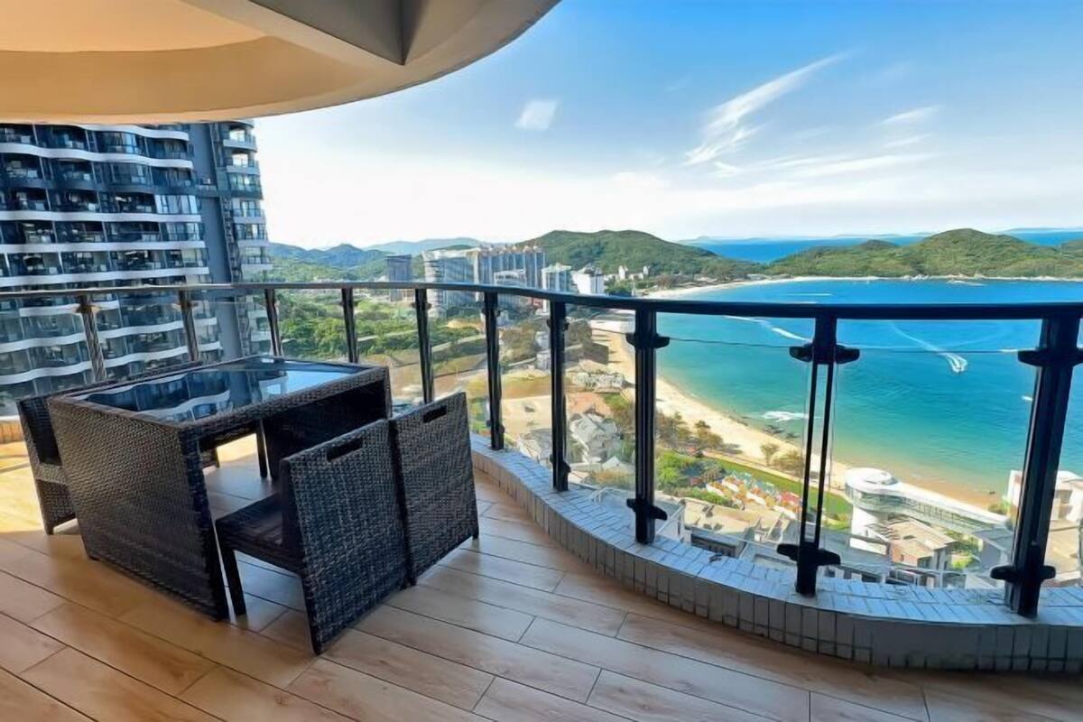 Un balcón con una mesa de vidrio y sillas de mimbre, con vista a una playa y un paisaje montañoso.