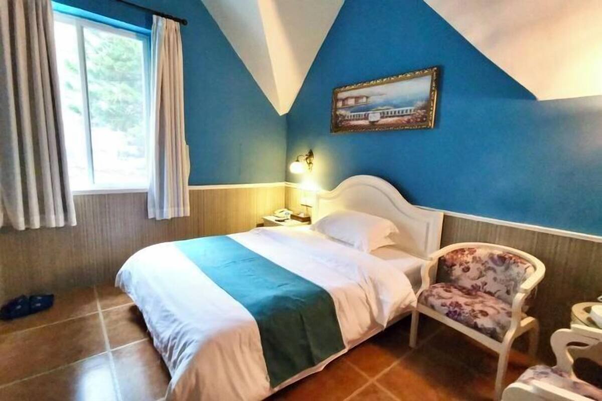 Un dormitorio con una cama, una silla, una mesita de noche, una lámpara, una ventana con cortinas y un cuadro en la pared.