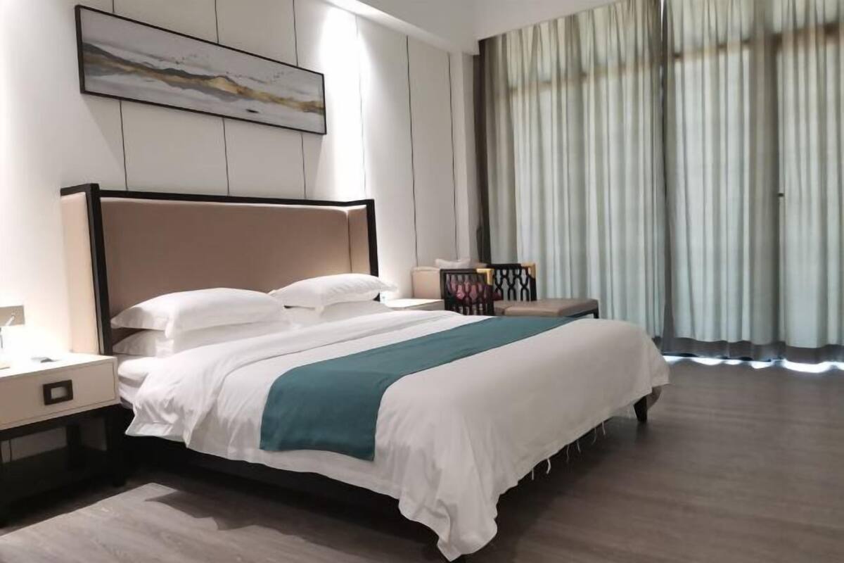 Una habitación de hotel moderna con una cama grande, una mesita de noche, una lámpara de noche, una obra de arte en la pared y una ventana con cortinas.