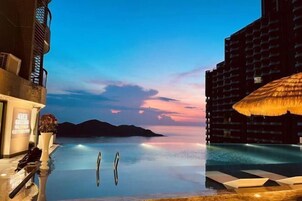 Un hotel con piscina infinita, una sombrilla de paja y vistas a montañas lejanas.