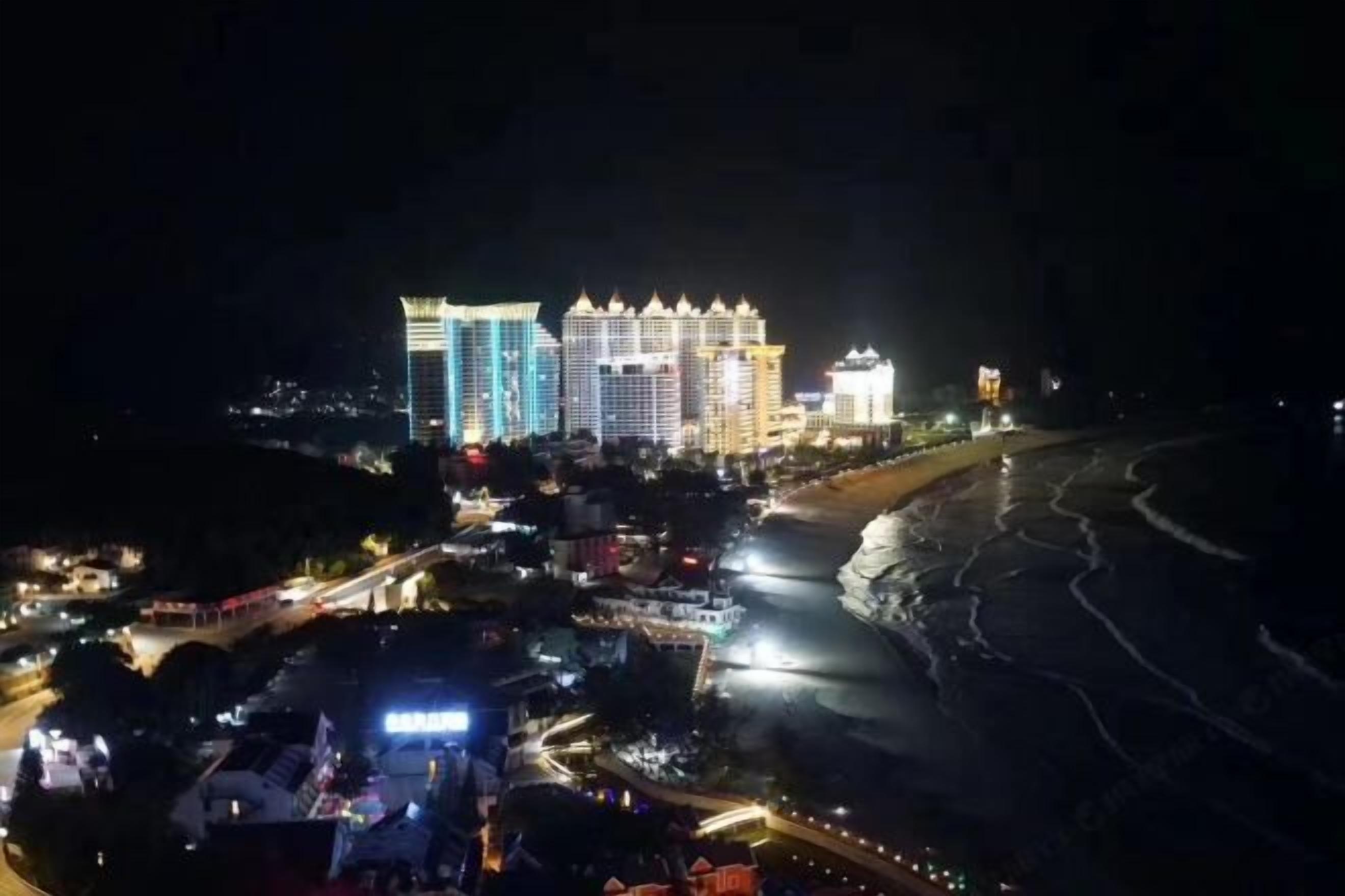 Un paisaje urbano costero nocturno con edificios iluminados, una carretera y una playa.