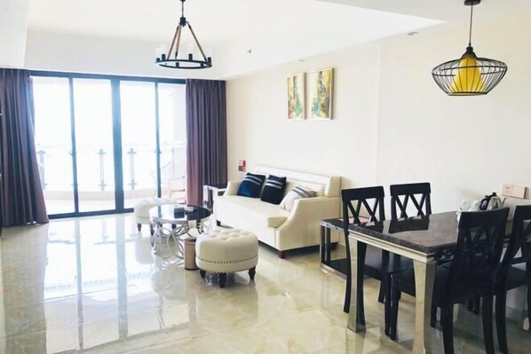 A modern living room with a sofa, dining table, and chairs. There is a large window with curtains, and artwork on the wall. - Peninsula Holiday Hotel (Nan'ao County)