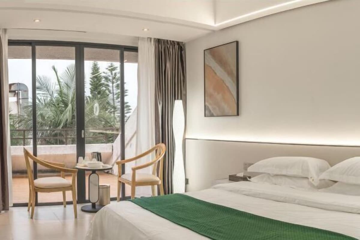 A hotel room with a bed, two chairs, a table, and a large window with a view of palm trees.