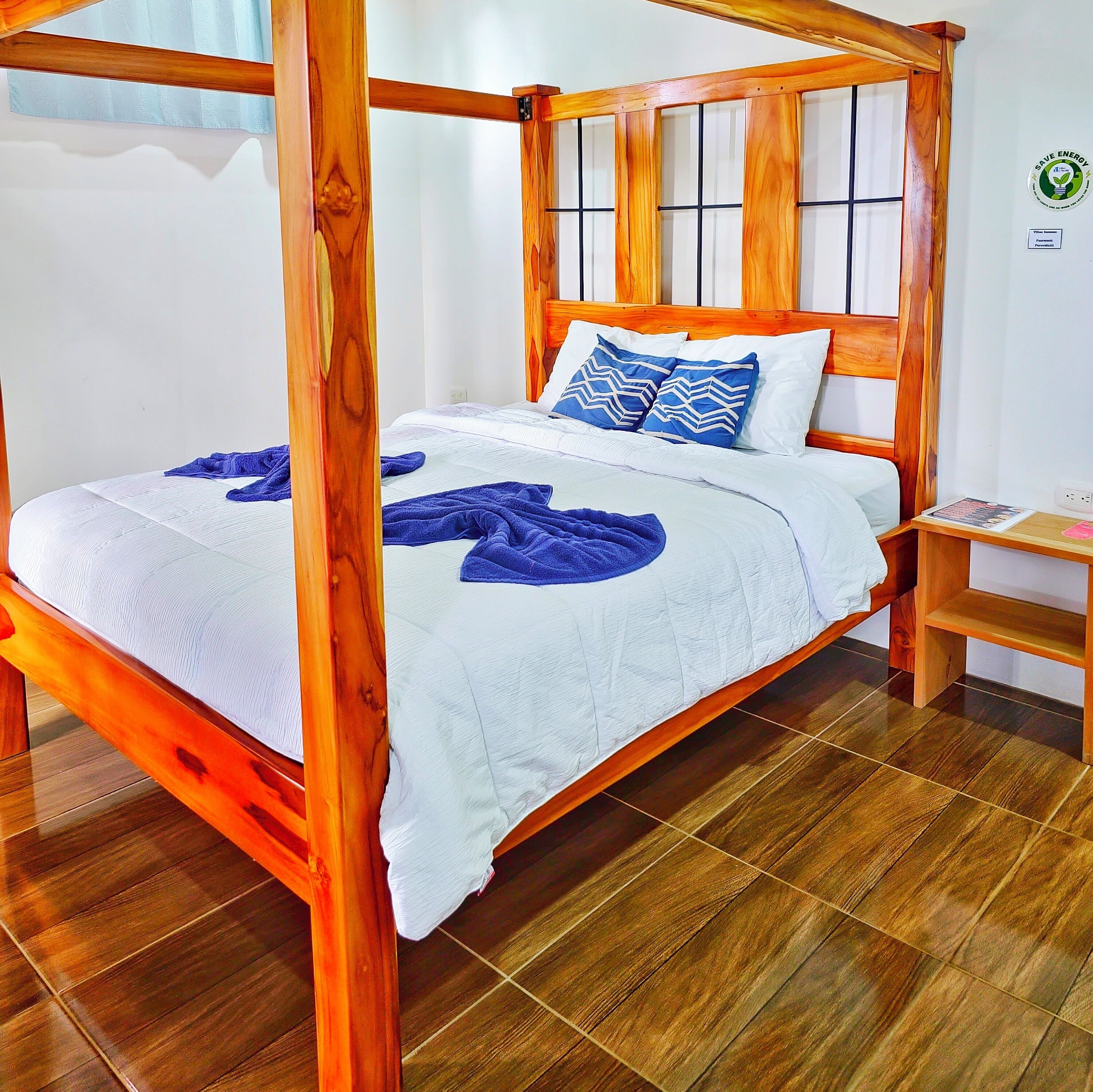 A wooden four-poster bed with a white quilt and blue accents, a wooden nightstand, and a tiled floor.