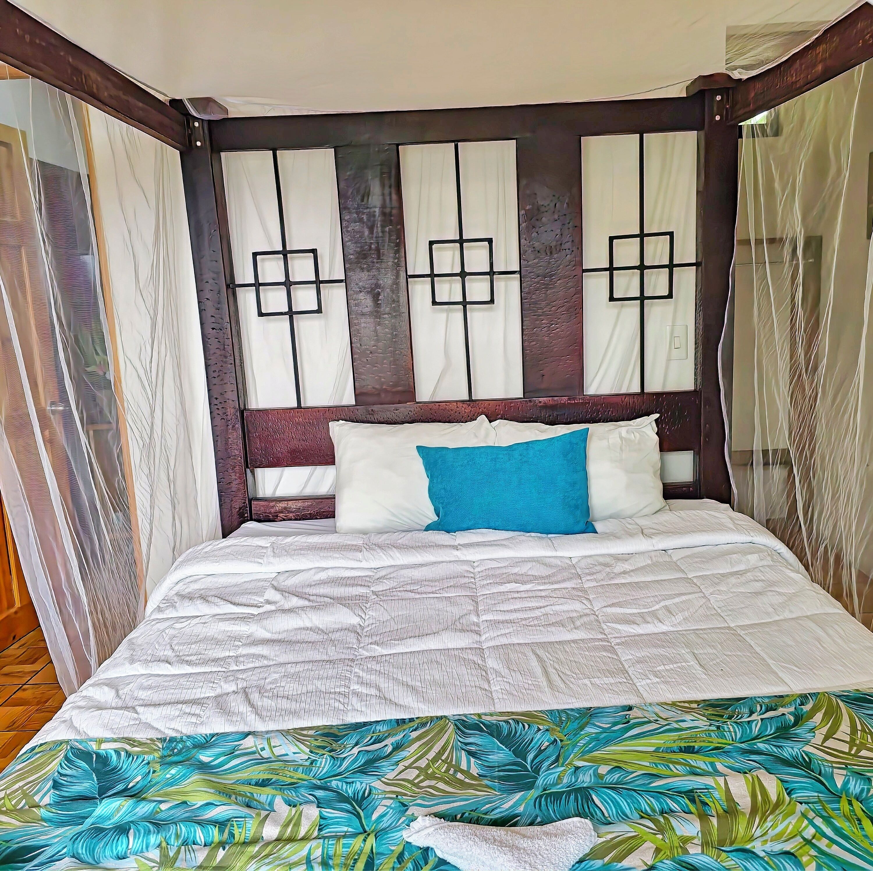 Una cama con una colcha blanca y azul estampada, un cabecero de madera con diseño geométrico y cortinas translúcidas en las ventanas.