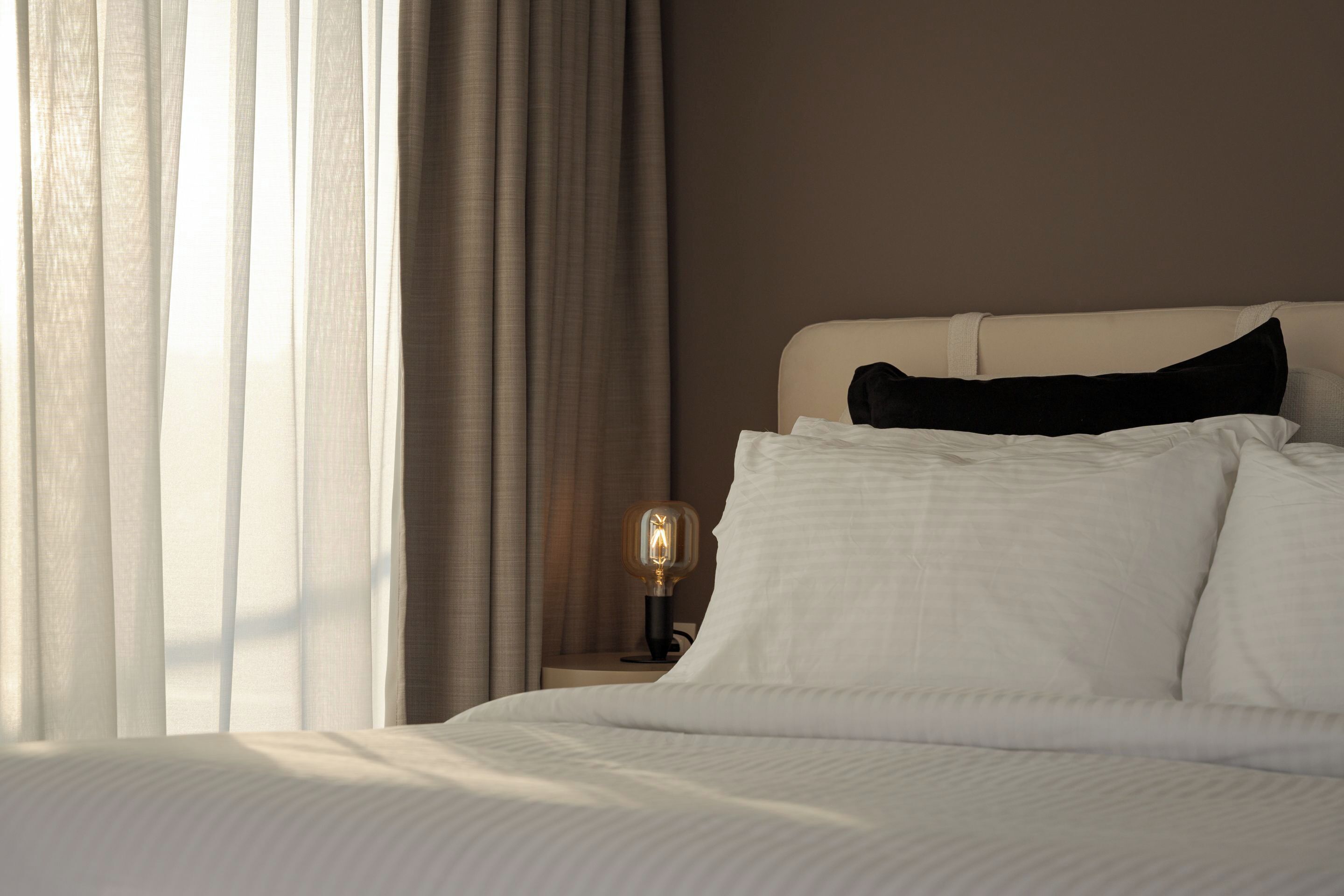 Een net opgemaakt bed met witte lakens en een zwart kussen, een bedlampje en een raam met dunne gordijnen.