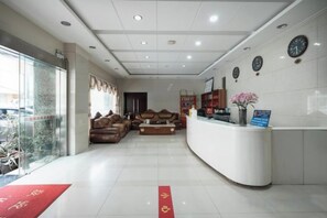 A reception area with a curved white reception desk, a sofa, and a coffee table. There are clocks on the wall and a window with a view of the outside. - Kunming Xiangyi Business Hotel (Kunming)