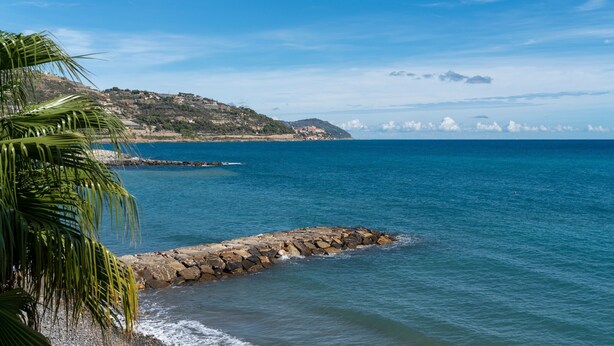 A coastal view with a rocky breakwater, clear blue sky, and distant hills.