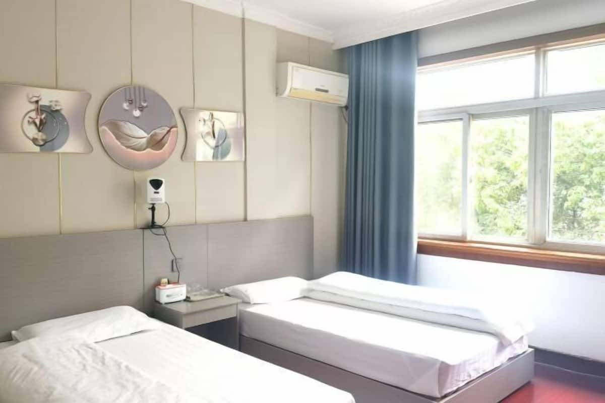 Habitación de hotel con dos camas, una mesita de noche, aire acondicionado instalado en la pared y una ventana con vista a zonas verdes.
