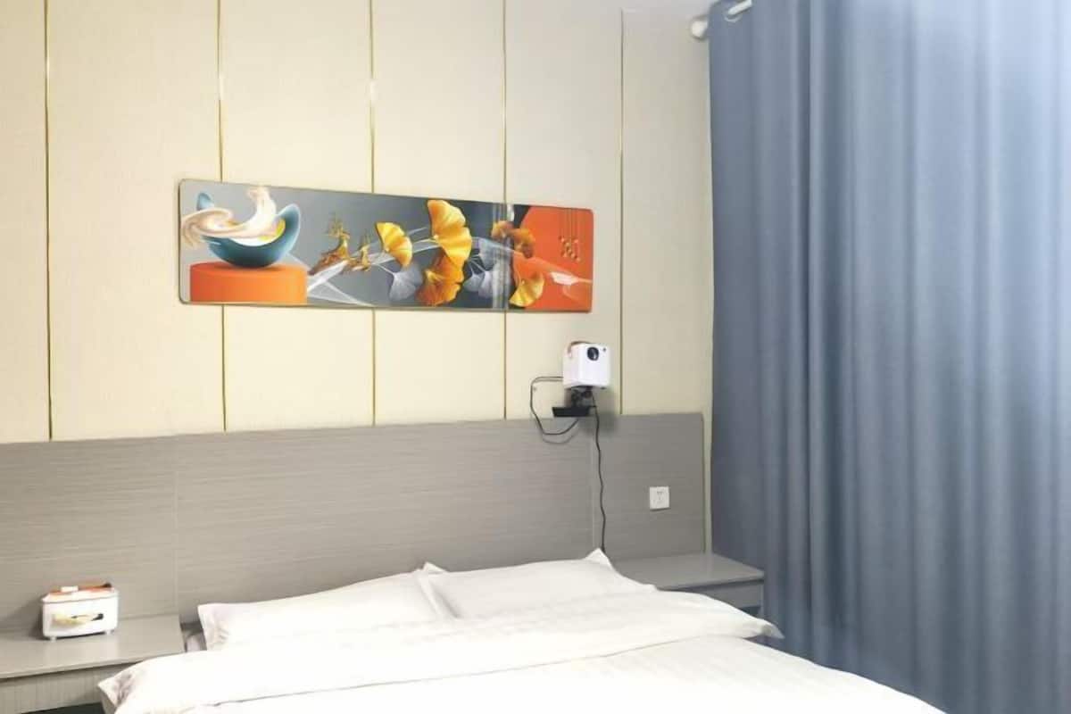 Una habitación de hotel con cama, mesita de noche, un cuadro, toma corriente y cortina.