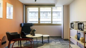 Una habitación con un ventanal grande, una silla negra, una mesita y un microondas en un estante.
