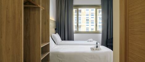 A hotel room with two beds, a wooden wardrobe, a window with curtains, and a view of buildings outside.