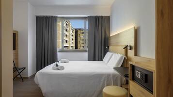 Quarto de hotel com cama, escrivaninha, cadeira, janela com cortinas e vista para edifícios.