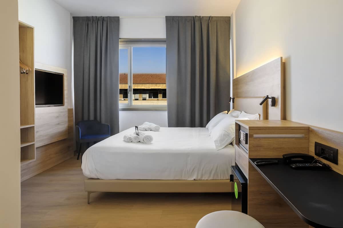 A modern hotel room with a bed, a desk, a chair, a TV, and a window with curtains.