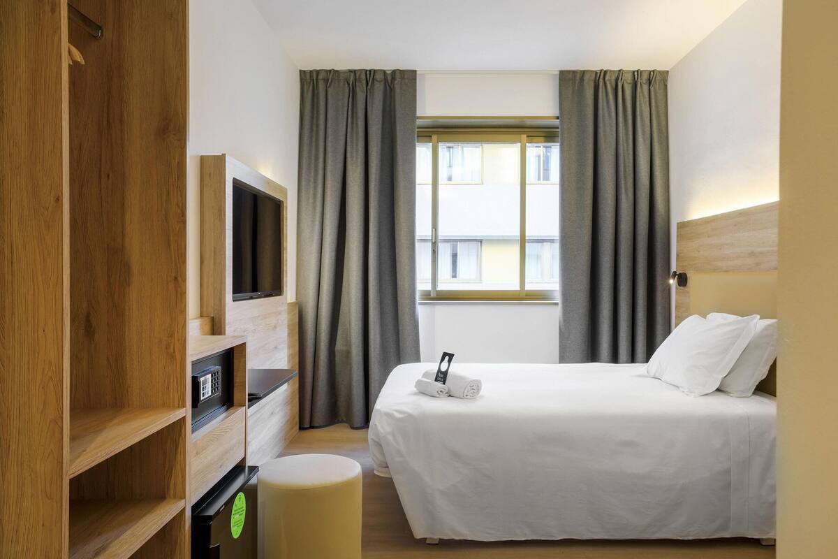 A hotel room with a bed, a TV, a wooden wardrobe, and a window with curtains.