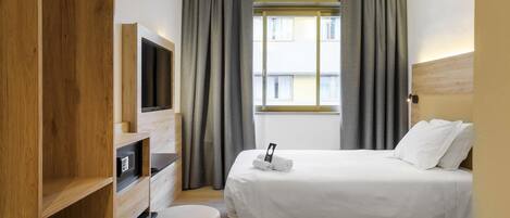 Ett hotellrum med en säng, en tv, en garderob i trä och ett fönster med gardiner.