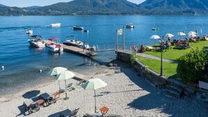 Ein See mit Strand, wo Boote liegen, ein Steg und eine Wiese mit Sonnenschirmen.