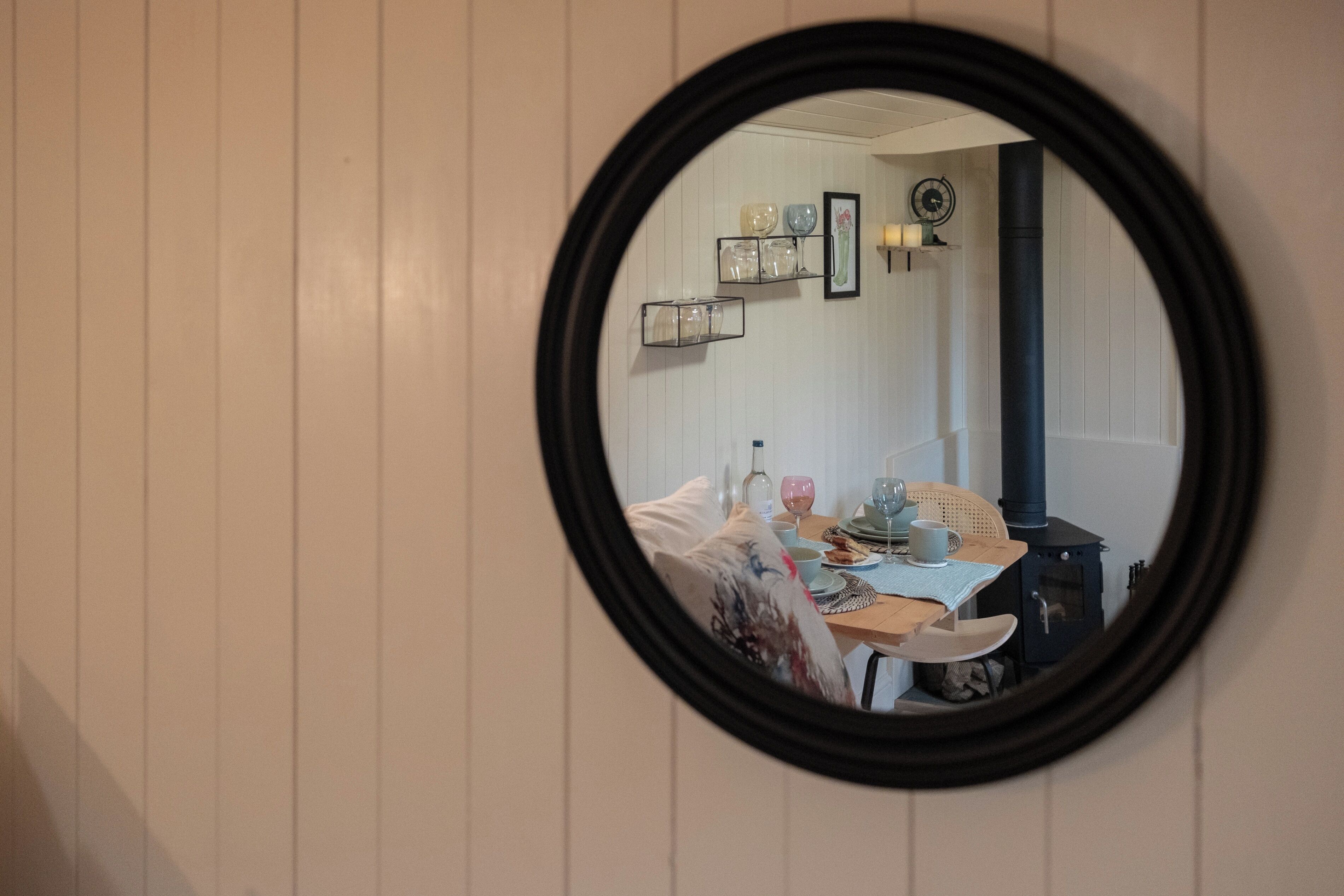 Un espejo redondo que refleja un comedor con una mesa preparada para dos, una silla y una estufa pequeña.