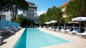 Une piscine rectangulaire à l’eau d’un bleu cristallin, entourée de chaises longues blanches et de parasols, avec un bâtiment visible au loin.