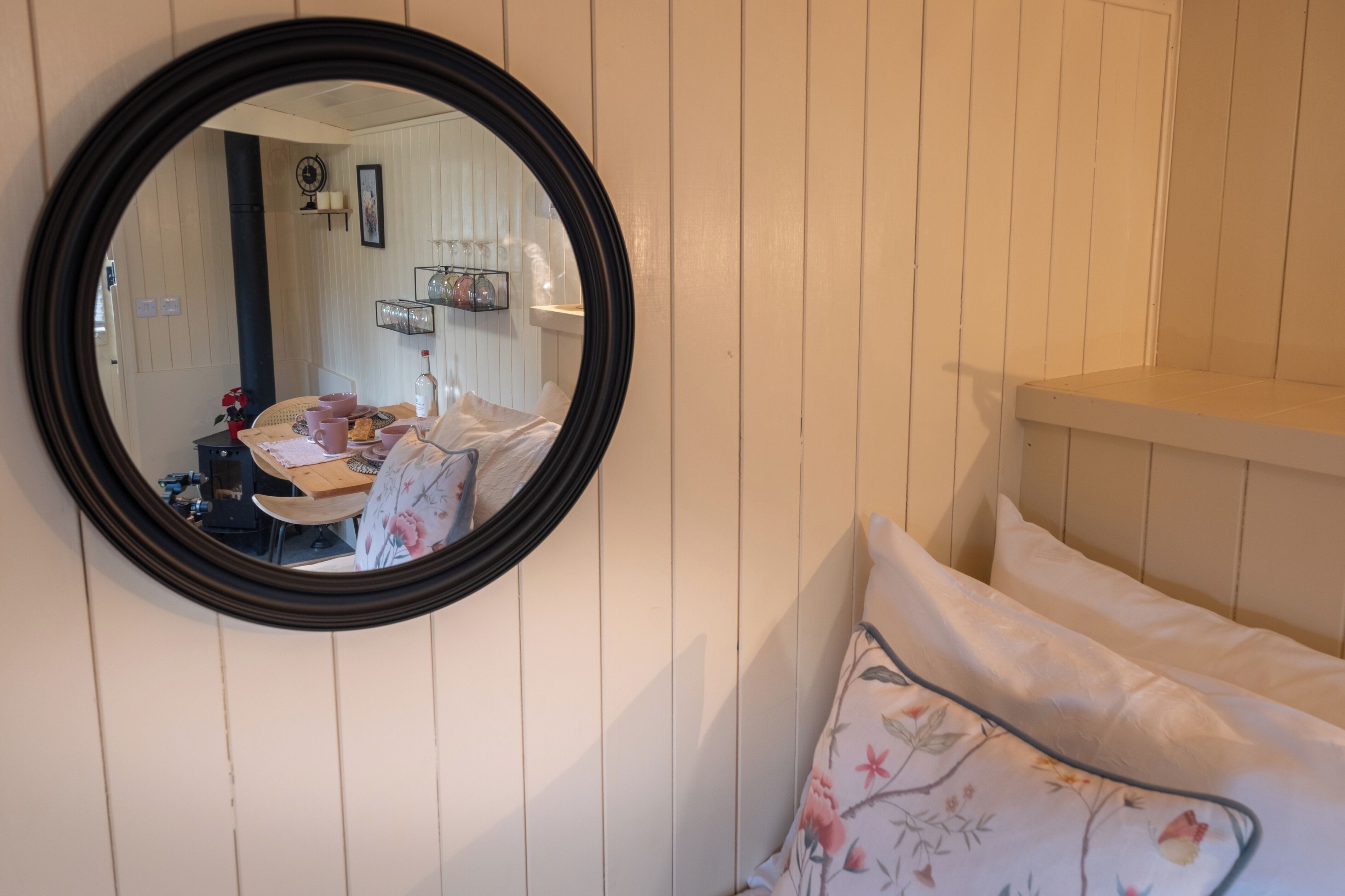 A room with a round mirror on the wall, a bed with floral bedding, and a small table with a chair.